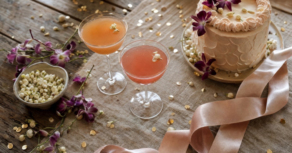 Overhead view of a bachelorette party table with cocktail glasses, tropical flowers, gold confetti, and a pastel cake in warm golden light