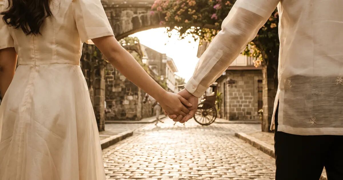 Couple holding hands while walking along cobblestone streets at golden hour, wearing cream dress and barong Tagalog, shot from behind with warm sunlight filtering through a stone archway