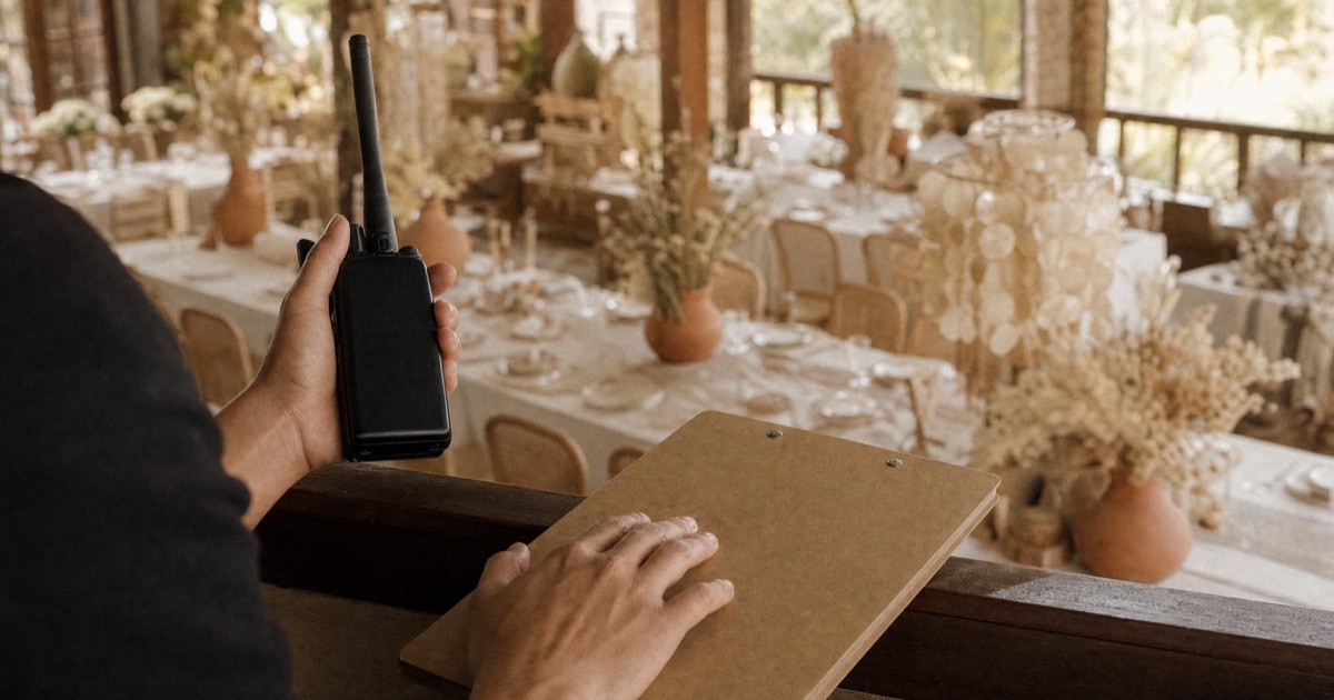 A wedding coordinator's hands on a two-way radio and a clipboard above a Filipino reception set with capiz lamps and dried flowers